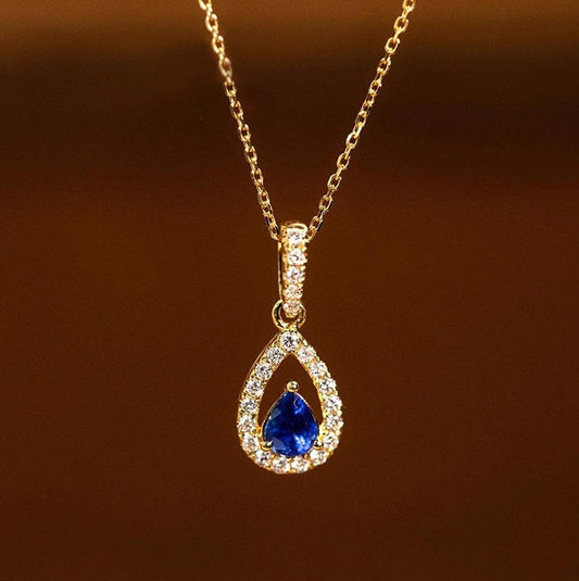 A gold teardrop pendant necklace featuring a central deep blue pear-cut gemstone surrounded by a halo of small clear stones, hanging against a warm brown gradient background.