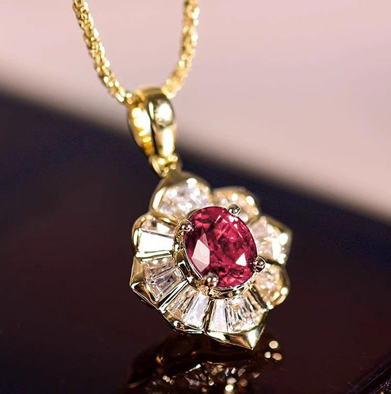 A gold pendant necklace featuring a central oval pinkish-red gemstone surrounded by clear baguette-cut stones, hanging from a gold chain against a dark reflective surface.