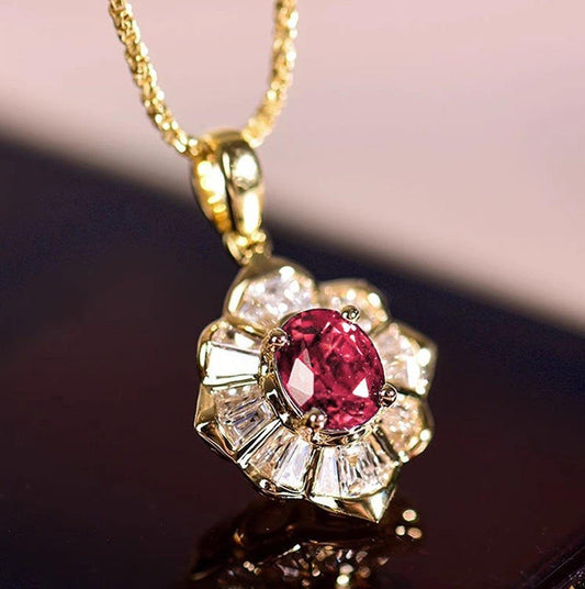 A gold pendant necklace featuring a central oval pinkish-red gemstone surrounded by clear baguette-cut stones, hanging from a gold chain against a dark reflective surface.