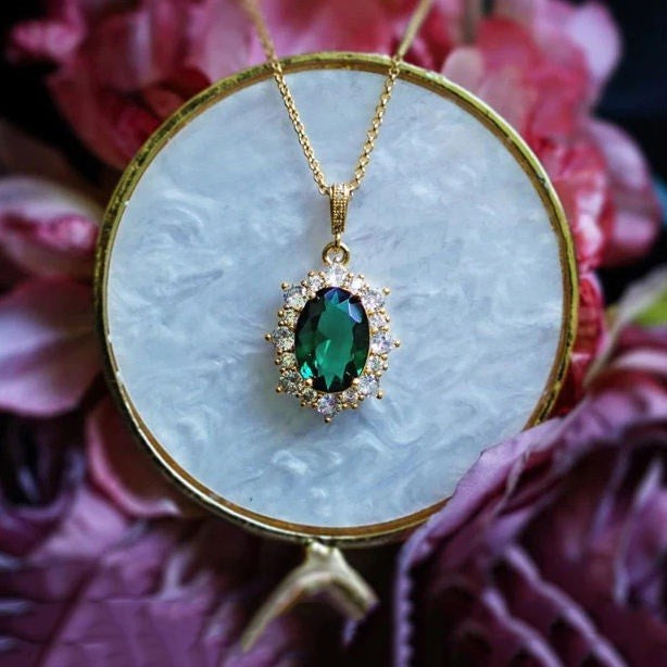 A gold pendant necklace with an oval green emerald-cut center stone surrounded by small clear stones, displayed on a round white stone surface with pink flowers in the background.