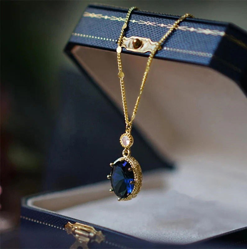 A gold pendant necklace with a large oval dark blue gemstone and a small clear accent stone, displayed hanging over an open dark blue jewelry box.