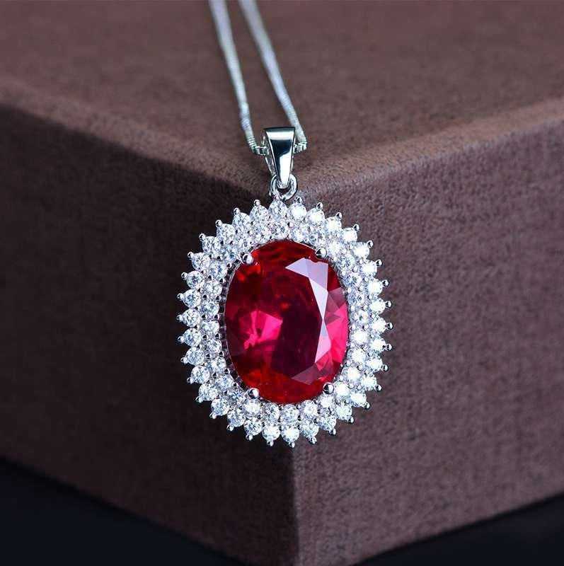 A silver pendant necklace featuring a large oval-cut red gemstone surrounded by a halo of small clear stones, displayed on a textured brown box.