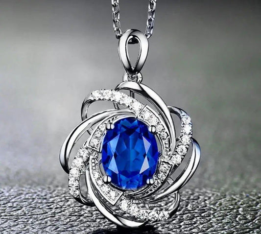 A silver-toned floral pendant necklace featuring a large oval-cut blue gemstone in the center, surrounded by smaller clear stones and curved silver bands, hanging from a chain against a dark gray background.