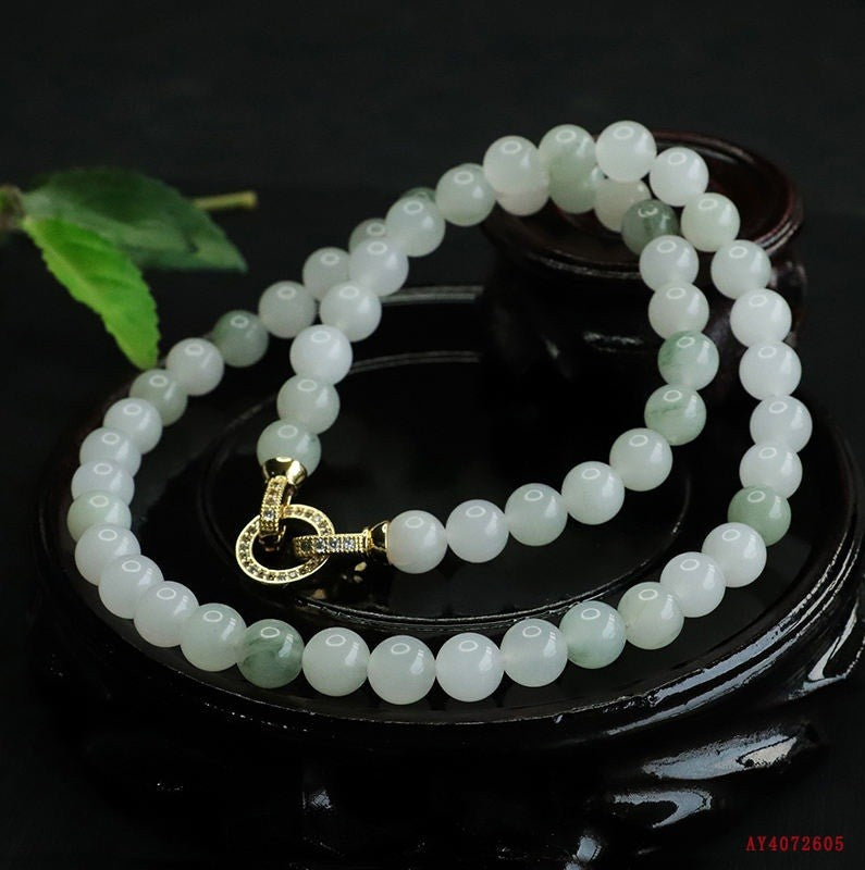 A beaded necklace made of pale green and white round beads with a gold clasp, displayed on a dark wooden stand against a black background.
