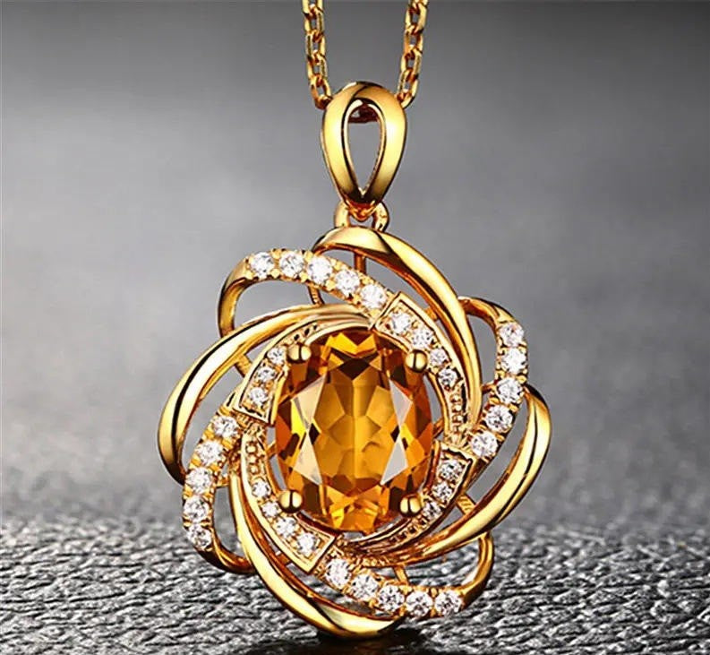 A gold pendant necklace featuring a large faceted orange-yellow center stone surrounded by swirling openwork gold bands set with small clear stones, shown on a dark textured background.