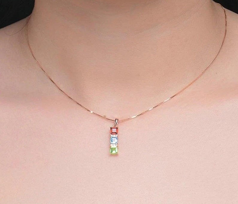 A close-up of a person's neck and upper chest wearing a delicate rose gold chain necklace with a vertical pendant featuring four small rectangular gemstones in red, blue, light blue, and green.