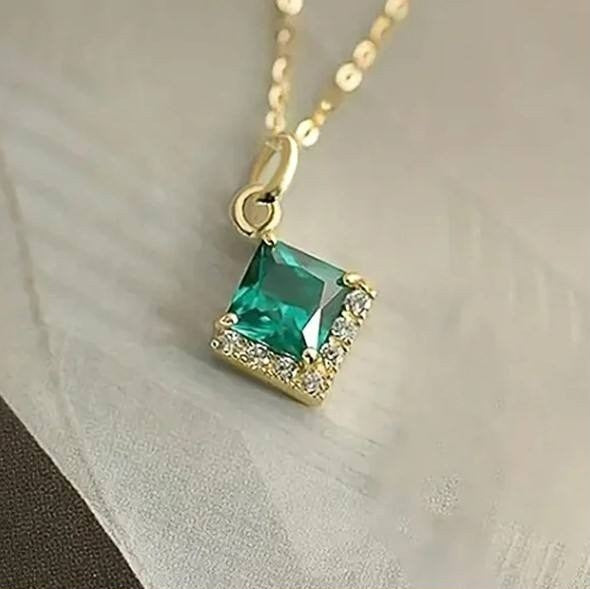 A gold pendant necklace featuring a square-cut green gemstone surrounded by small clear crystals, resting on a light textured fabric surface.
