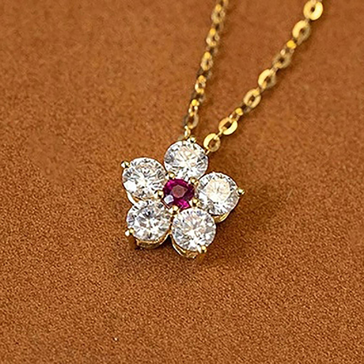 A gold flower-shaped pendant necklace featuring five clear round-cut stones surrounding a small red center stone, displayed on a textured brown background.