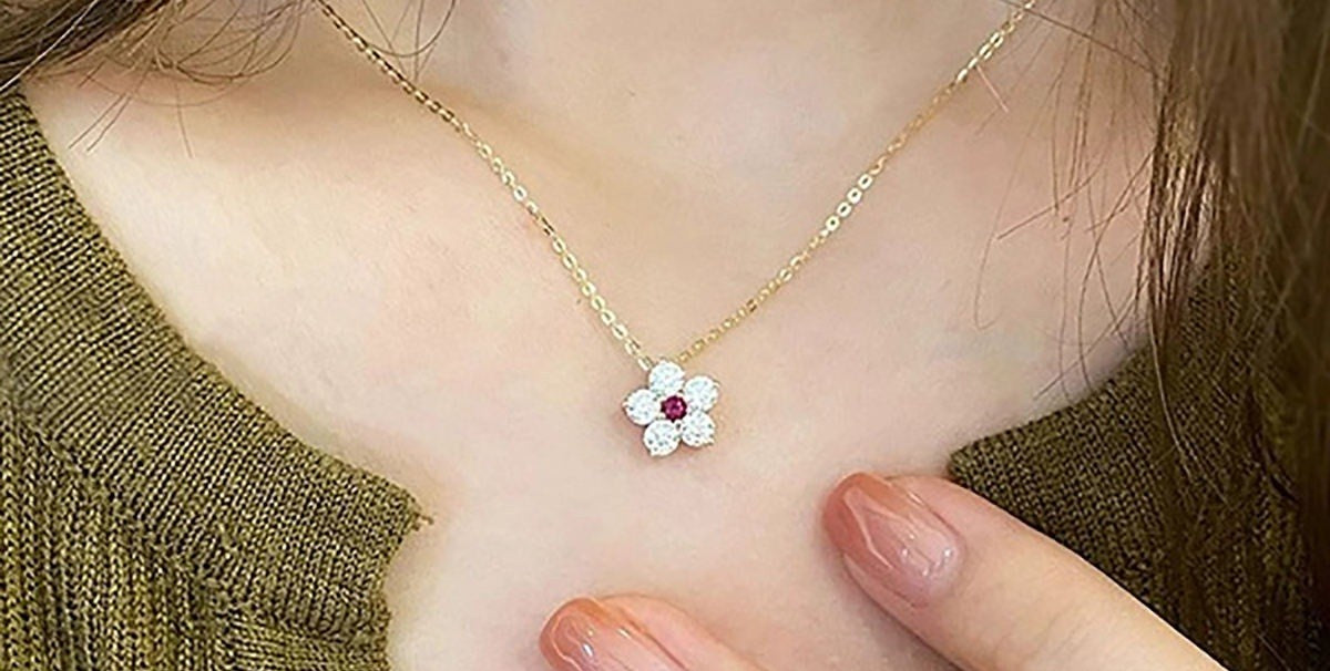 A close-up of a person wearing a delicate gold chain necklace with a small flower-shaped pendant. The pendant features clear rhinestones in its petals and a small red gemstone in the center, worn against a green knit top.