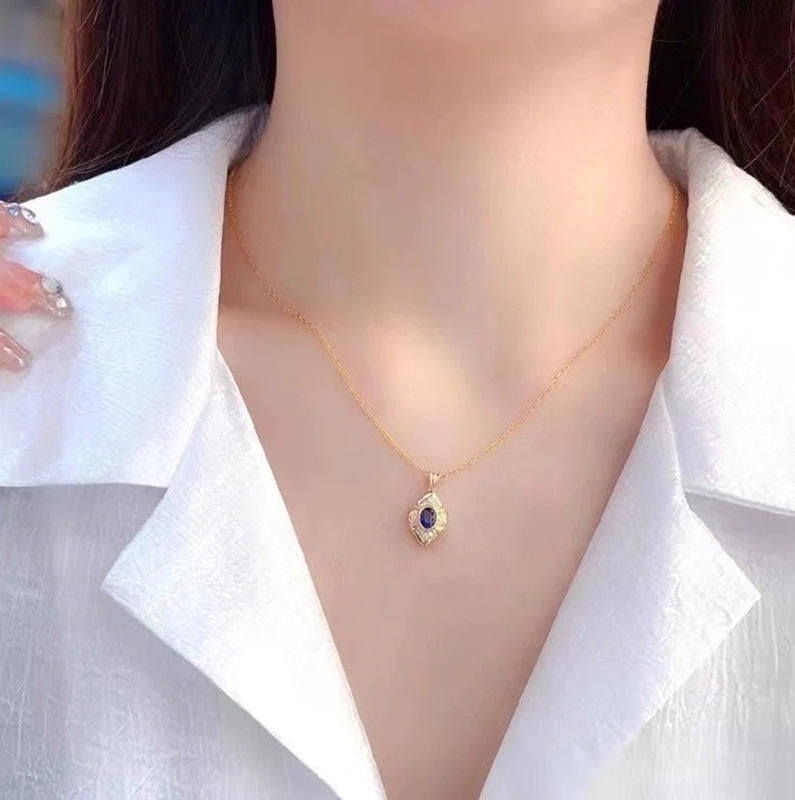 A close-up of a woman wearing a delicate gold chain necklace with a teardrop-shaped pendant featuring a dark blue center stone and small clear stones around it, styled with a white collared shirt.