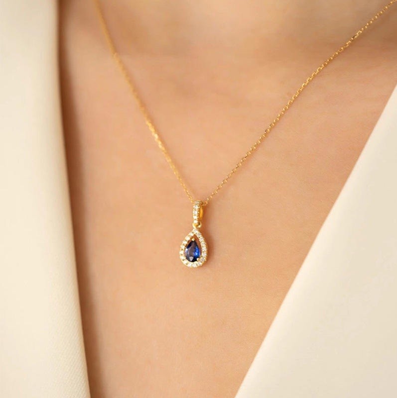 A gold teardrop pendant necklace with a deep blue sapphire center stone and small clear accent stones, worn on a model's neck over a cream-colored top.