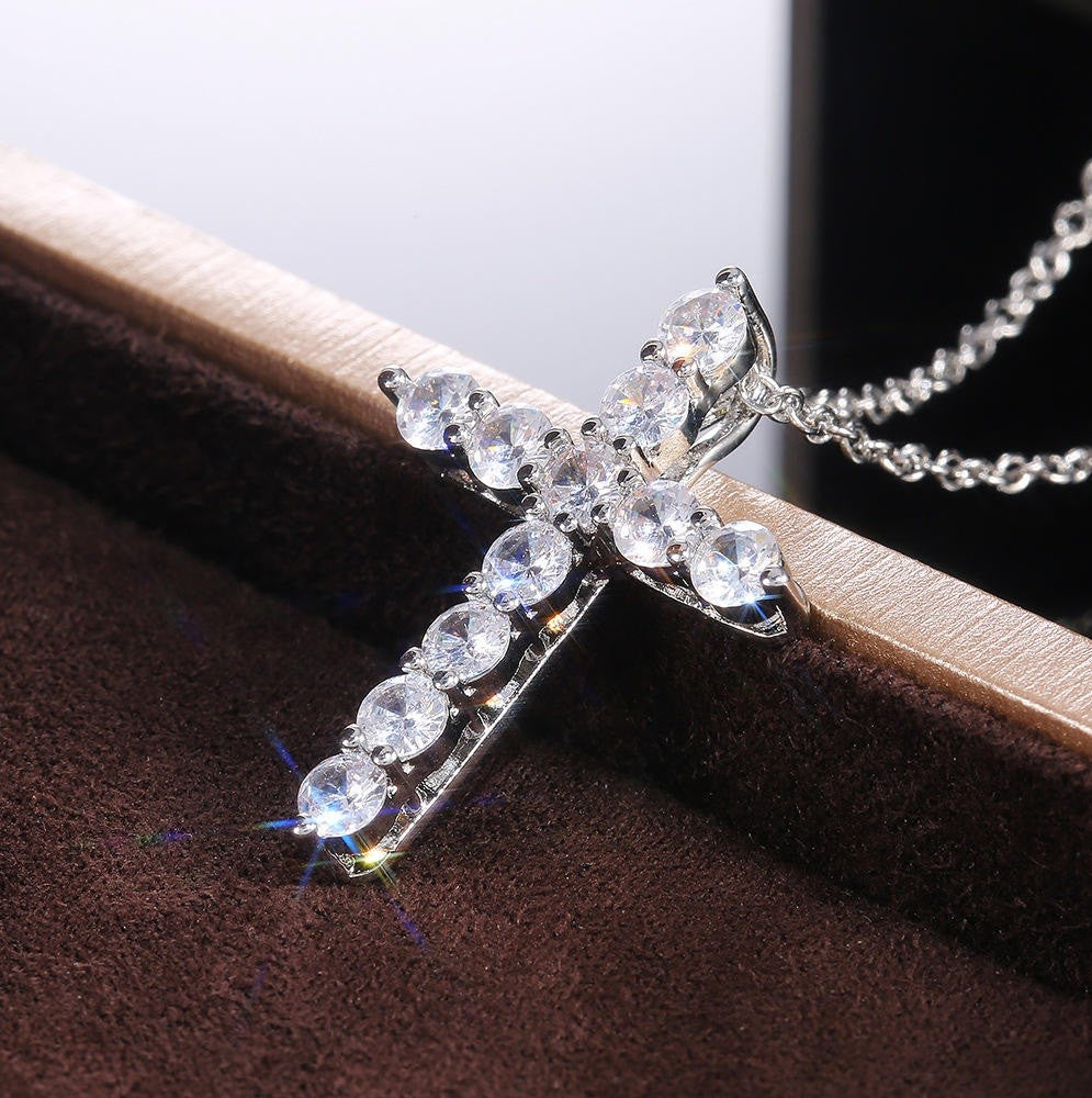 A silver-toned cross pendant necklace adorned with clear round-cut rhinestones, resting on a brown velvet jewelry box with a white background visible behind it.