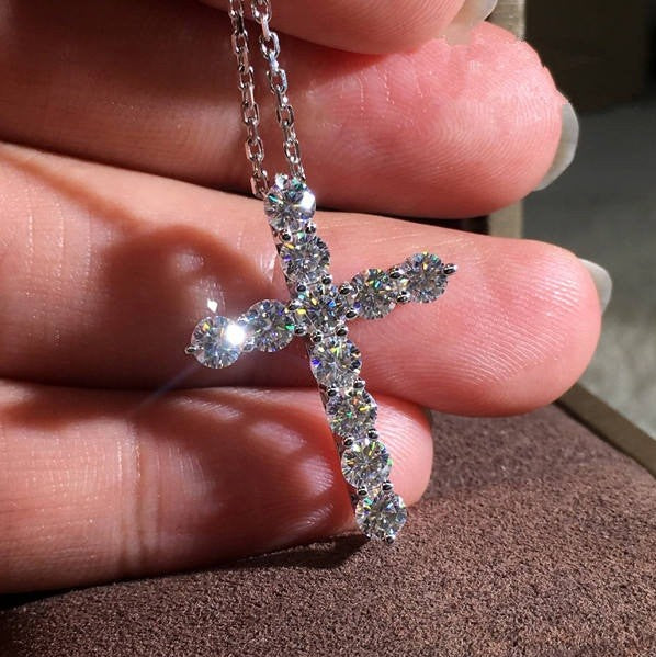 Silver cross pendant necklace held between fingers on a textured surface