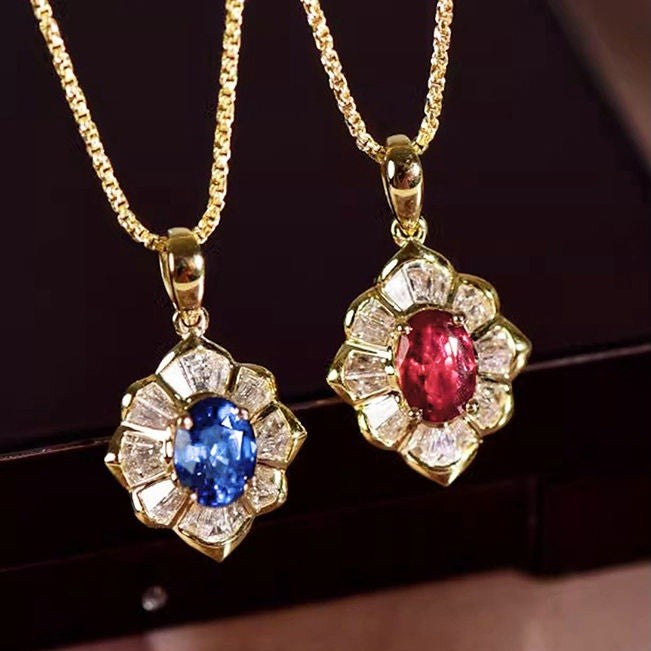 Two gold pendant necklaces on chains, each featuring a flower-shaped setting with clear stones and a central oval gemstone, one blue and one red, displayed against a dark background.