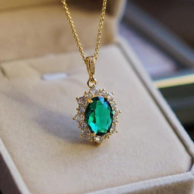 A gold pendant necklace with a large oval emerald-cut green gemstone surrounded by small clear stones, displayed on a beige jewelry box.