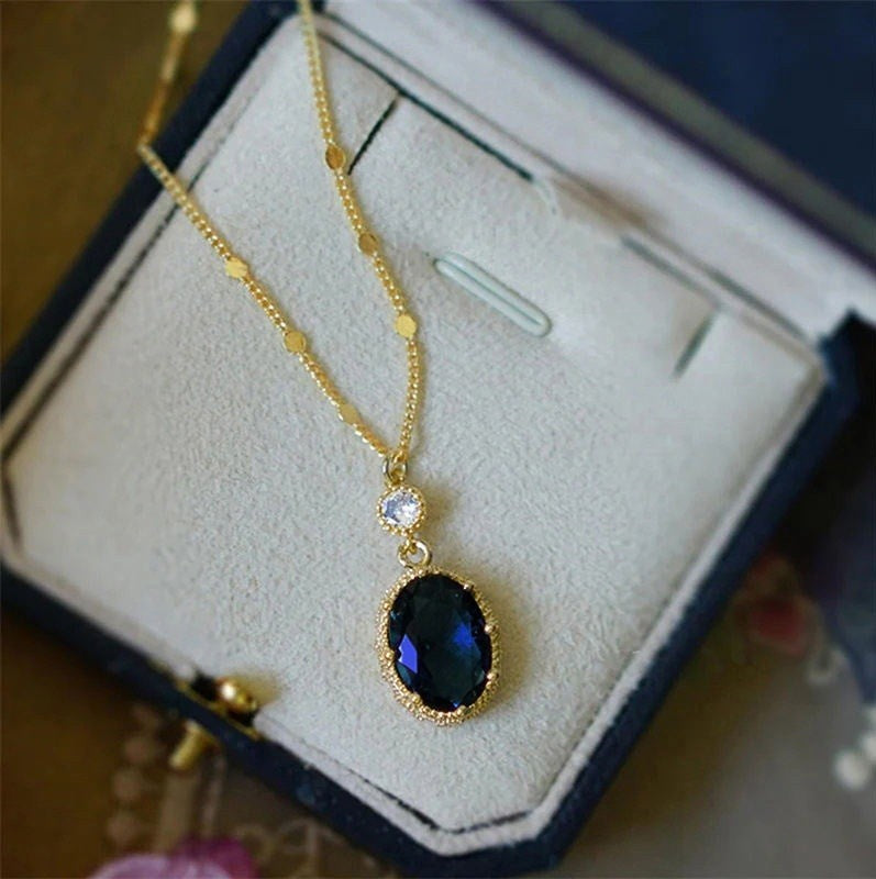A gold necklace with an oval dark blue gemstone pendant and a small clear accent stone, displayed inside an open jewelry box.