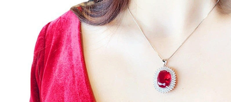 A close-up of a person wearing a silver-toned pendant necklace with a large oval red gemstone, paired with a red top against a white background.