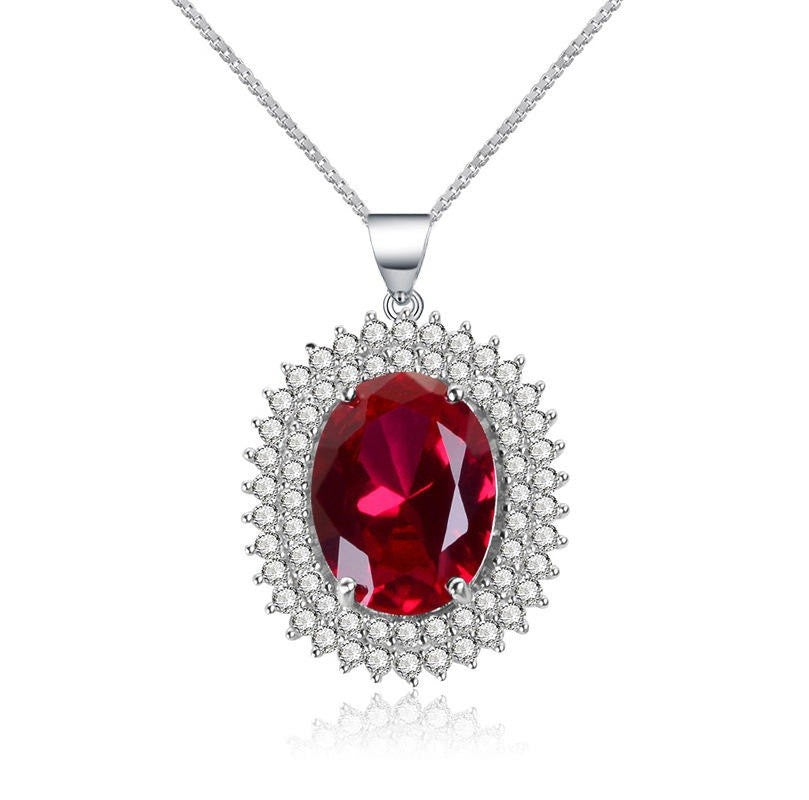 A silver pendant necklace featuring a large oval-cut red gemstone surrounded by a halo of small clear stones, suspended from a silver chain on a white background.