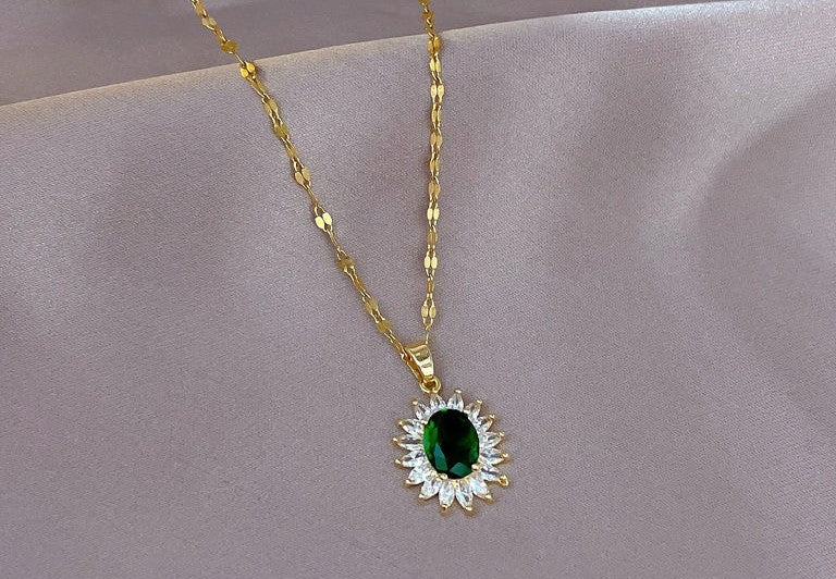 A gold chain necklace with a sunburst pendant featuring a central green oval stone surrounded by small clear stones, displayed on a soft beige fabric background.