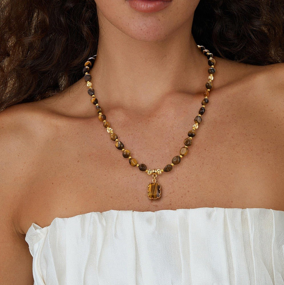 A close-up of a woman wearing a beaded necklace with a tiger's eye pendant. The necklace features faceted beads in brown, black, and gold, with a polished gold-toned pendant.