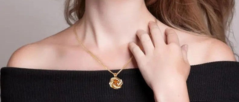 A close-up lifestyle shot of a model wearing a delicate gold necklace with a rose-shaped pendant containing amber-colored stones, styled with a black off-the-shoulder top.