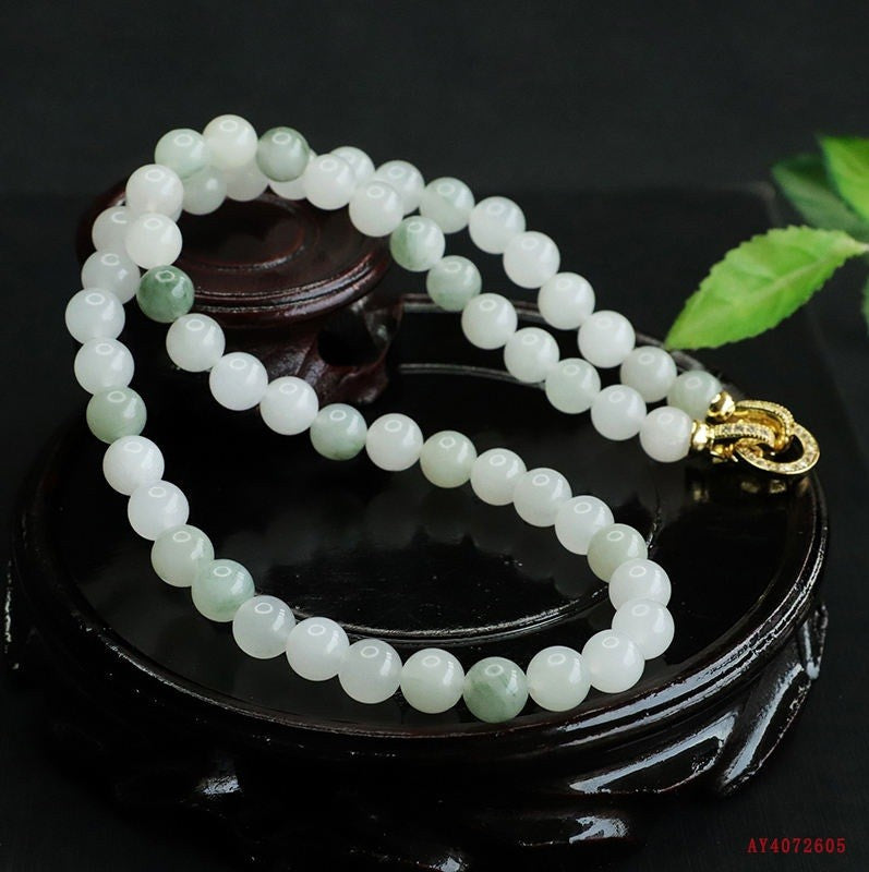 A beaded necklace made of round white and pale green jade-like beads with a gold clasp, displayed on a dark wooden stand against a black background.