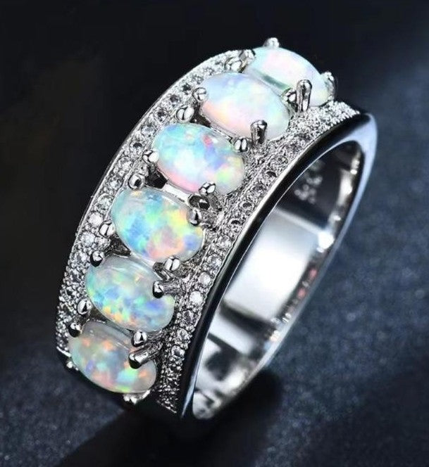 A wide silver band ring featuring five oval opal gemstones with iridescent rainbow play-of-color, surrounded by pavé-set clear stones, photographed on a dark textured background.
