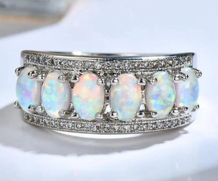A silver band ring featuring five oval opal stones with iridescent white, blue, and pink flashes, surrounded by pavé-set clear stones on a white background.