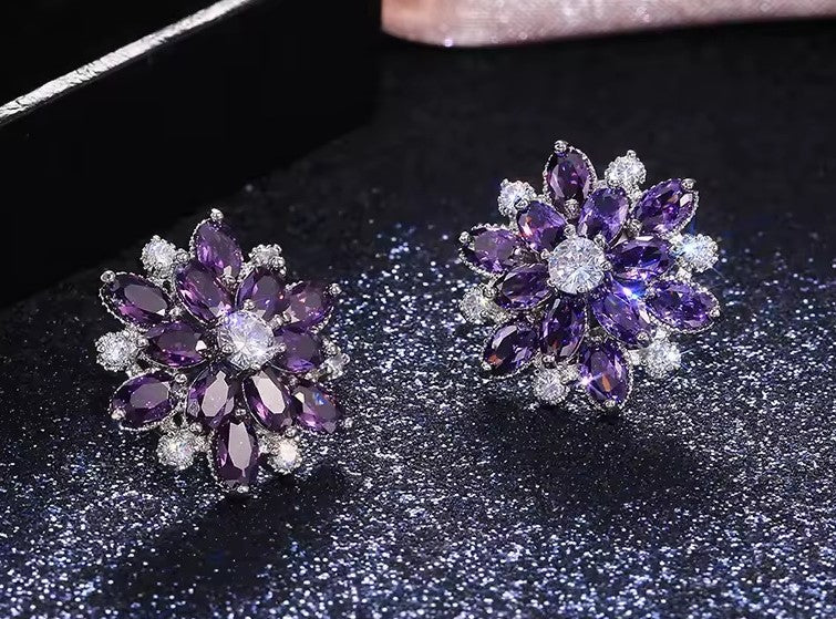 A pair of flower-shaped stud earrings with deep purple gemstones and clear accent stones, displayed on a dark glittery surface.