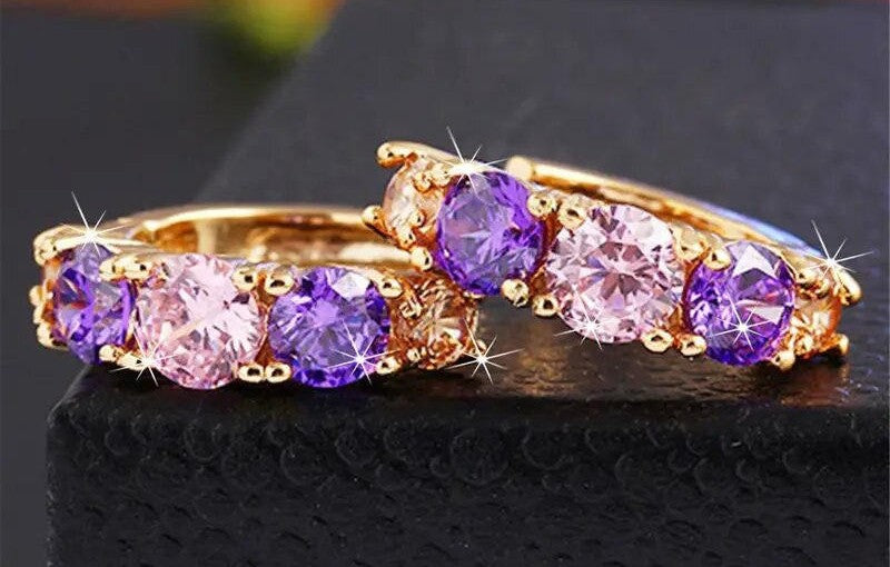 A pair of gold rings featuring round-cut purple and light pink gemstones, displayed on a textured black surface.