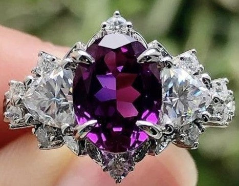 A close-up of a silver ring featuring a large oval purple gemstone in the center, flanked by clear round and marquise-cut gemstones, held against a blurred green background.