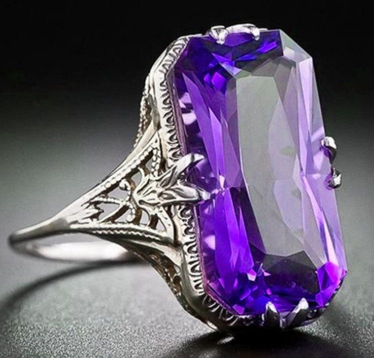 A silver ring featuring a large, faceted rectangular purple gemstone, photographed against a dark, reflective background.