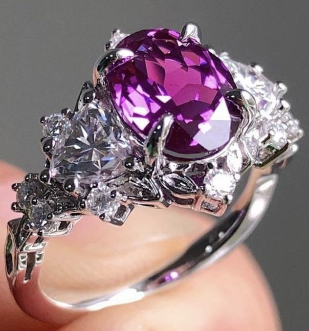 A silver ring with a large oval-cut purple gemstone and clear accent stones, shown being worn on a finger against a neutral gray background.