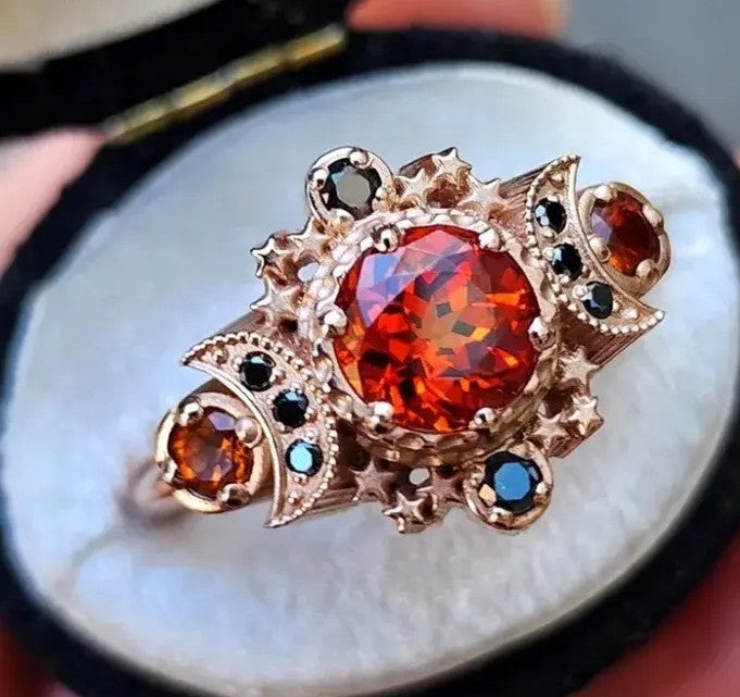 A close-up of a vintage-style rose gold ring with a large round orange-red central gemstone and smaller orange, black, and blue accent stones, displayed on a white ring box.