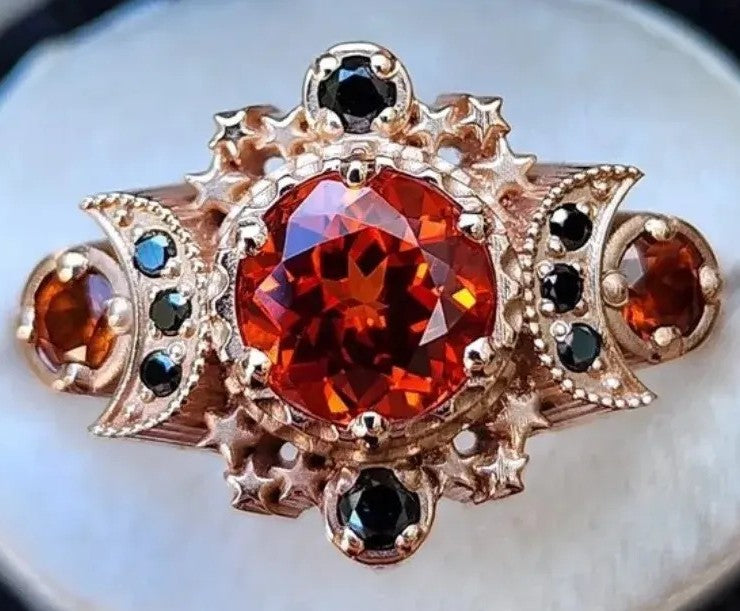 A vintage-style gothic ring featuring a large round orange-red center gemstone, accented by smaller black and blue stones in a rose gold setting.