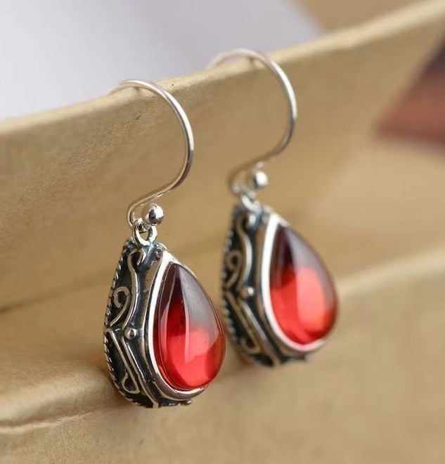 A pair of silver-toned drop earrings with ornate filigree sides and teardrop-shaped red gemstones, displayed on a beige surface.