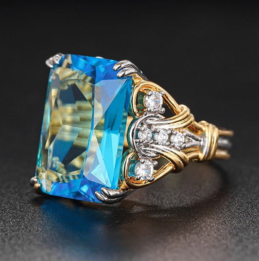 A close-up shot of a ring featuring a large rectangular blue gemstone in a yellow gold and silver setting with small clear accent stones, set against a dark background.