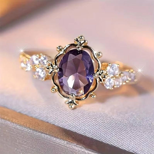 A gold ring featuring a large oval purple gemstone in the center, flanked by clear accent stones on a soft pink background.
