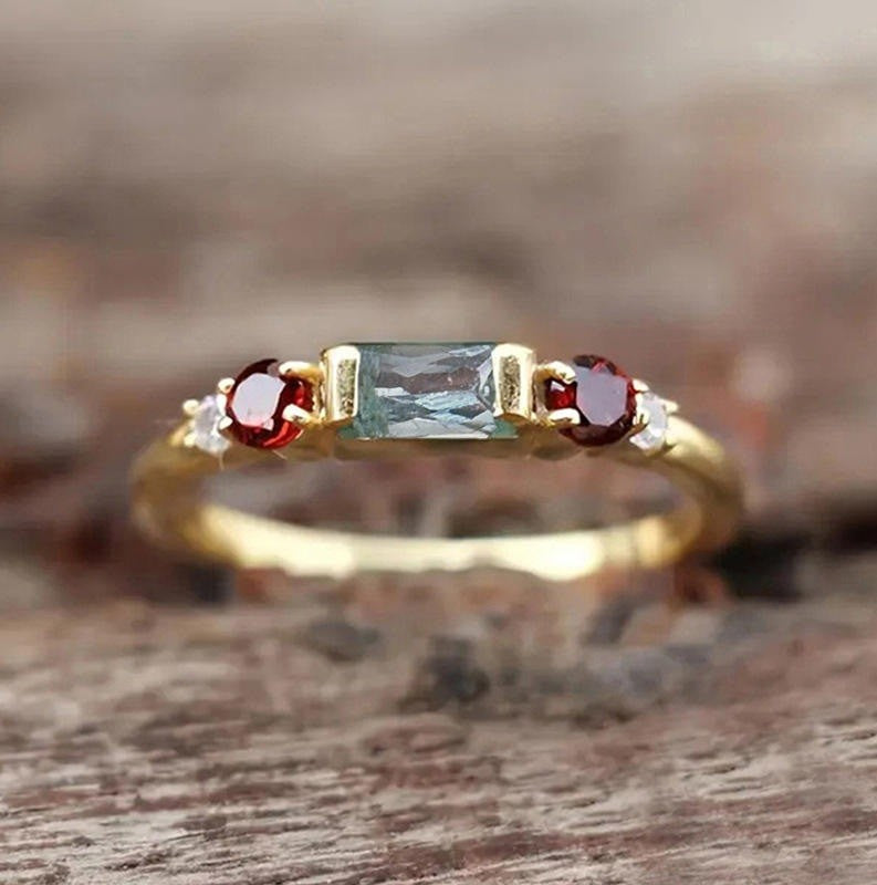 A yellow gold three-stone ring featuring a rectangular emerald-cut light blue center stone, flanked by round red garnet and small clear accent stones, resting on a rustic wooden surface.