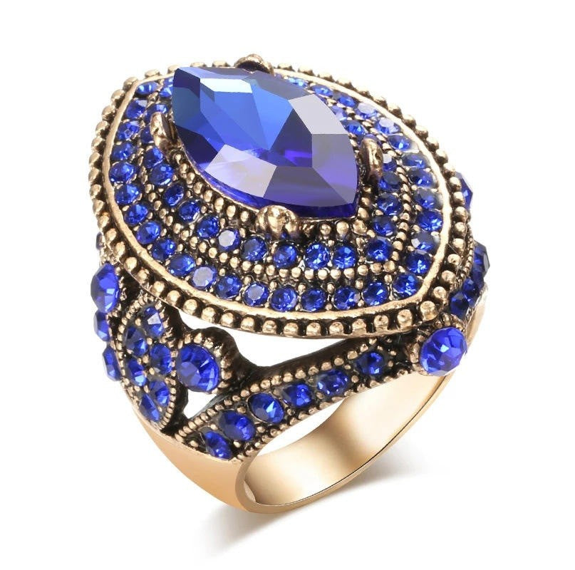 A statement ring featuring a large marquise-cut deep blue crystal surrounded by multiple tiers of small round blue crystals in an antique gold-tone setting, shown on a clean white background.