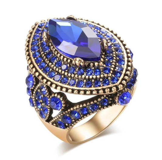 A statement ring featuring a large marquise-cut deep blue crystal surrounded by multiple tiers of small round blue crystals in an antique gold-tone setting, shown on a clean white background.