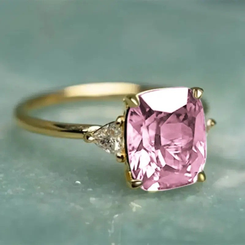 A yellow gold ring featuring a large cushion-cut pink gemstone and a small round diamond accent on the band, set against a soft, light blue-green background.