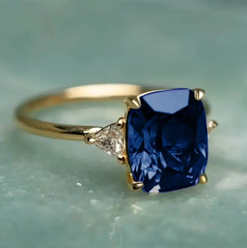 A yellow gold ring featuring a large cushion-cut deep blue sapphire center stone and a small round diamond accent on the band, set against a soft, pale green textured background.