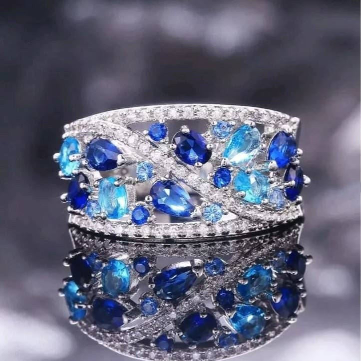 A wide silver-toned band ring embellished with marquise-cut and round deep blue sapphires and small clear stones, shown against a dark blurred background with a mirrored reflection below.