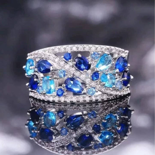 A wide silver-toned band ring embellished with marquise-cut and round deep blue sapphires and small clear stones, shown against a dark blurred background with a mirrored reflection below.