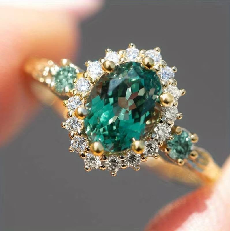 A yellow gold ring with a large oval green emerald-cut center stone and surrounding clear accent stones, shown being held between fingers against a soft gray background.