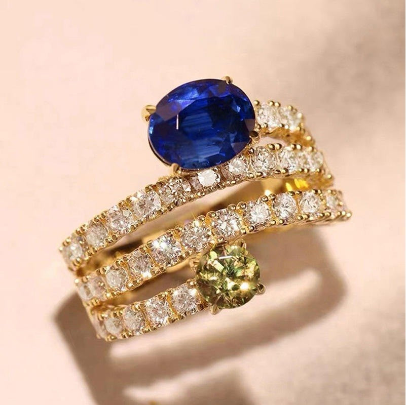 A gold ring set featuring a large oval-cut blue sapphire, a round-cut yellow-green peridot, and multiple bands paved with clear diamonds, displayed on a soft beige background.