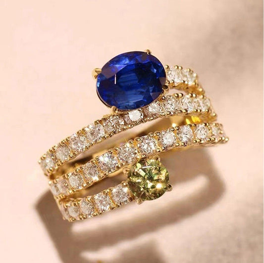 A gold ring set featuring a large oval-cut blue sapphire, a round-cut yellow-green peridot, and multiple bands paved with clear diamonds, displayed on a soft beige background.