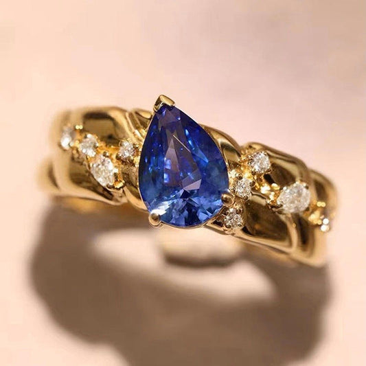 Gold ring with a blue pear-shaped gemstone and smaller diamonds on a beige background