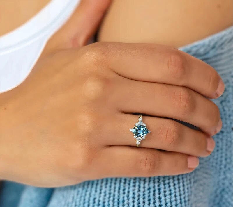 A close-up of a person's hand wearing a silver ring with a round-cut light blue gemstone, resting on a blue knit sweater.